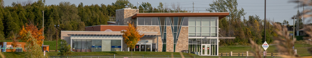 A modern building with large glass windows and a mix of brick and metal exterior, surrounded by greenery including trees with autumn-colored leaves. There is a sign in front of the building, but the text is not clearly visible. The sky is clear, indicating good weather.